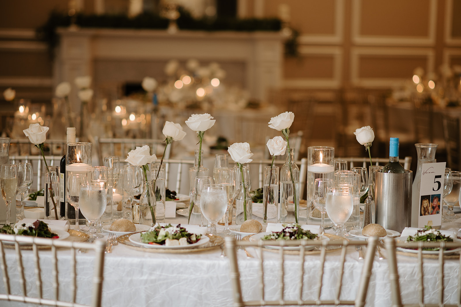 Reception table setting featuring bud vases with white roses, warm candles, and plated salads.